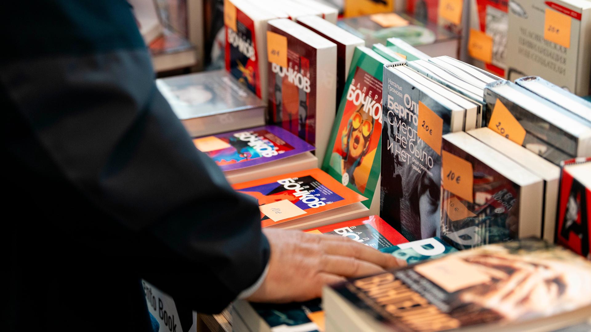 Auf der russischsprachigen Buchmesse in Berlin werden verschiedene russischsprachige Bücher zum Verkauf angeboten. Abgebildet ist ein Tisch, auf dem verschiedene Bücher aufgestellt und gestapelt liegen. Die Hand eines Mannes berührt eines der Bücher. Auf der russischsprachigen Buchmesse in Berlin werden verschiedene russischsprachige Bücher zum Verkauf angeboten. Abgebildet ist ein Tisch, auf dem verschiedene Bücher aufgestellt und gestapelt liegen. Die Hand eines Mannes berührt eines der Bücher.