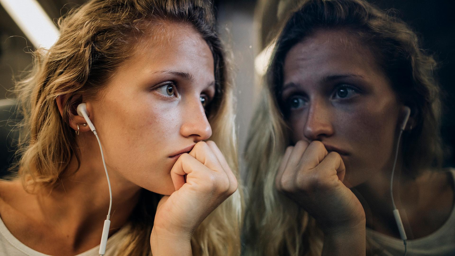 Eine junge Frau mit blonden Locken sitzt mit Kopfhörern am Zugfenster und schaut traurig aus dem Fenster, in dem sich ihr Gesicht spiegelt. Eine junge Frau mit blonden Locken sitzt mit Kopfhörern am Zugfenster und schaut traurig aus dem Fenster, in dem sich ihr Gesicht spiegelt.