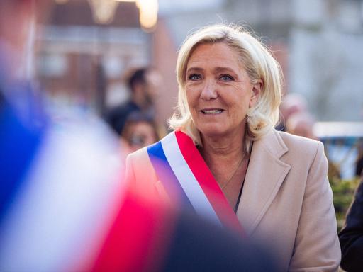 Marine Le Pen trägt ein helles Sakko und eine Schärpe mit den französischen Nationalfarben. Im Vordergrund, in der Unschärfe ist eine weitere Person mit Schärpe zu erkennen.