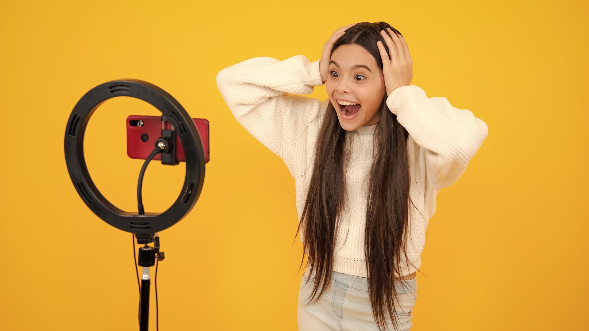 Eine junge Bloggerin vor dem Kameraphone mit weit aufgerissenen Augen vor gelbem Hintergrund.