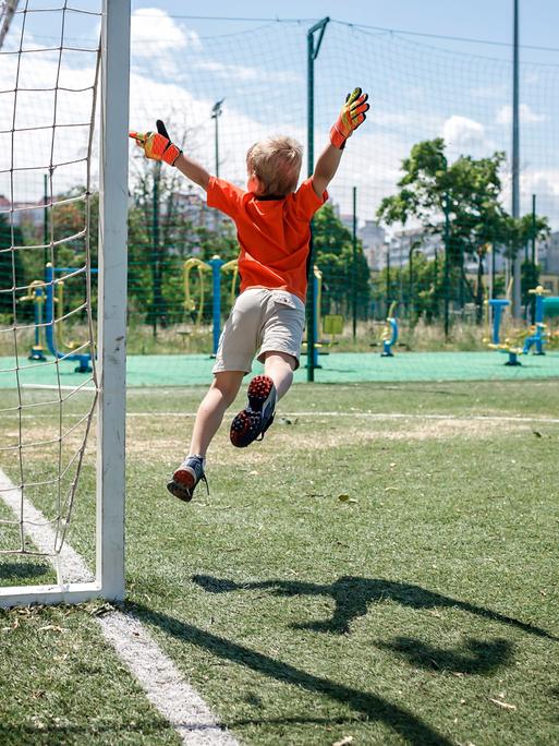 Ein Kind in Fußballkleidung beim Sprung durch die Luft vor einem Tor.