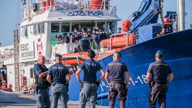 Auf einem Schiff im Hafen von Lampedusa warten viele Menschen darauf, an Land gehen zu können. Vor dem Schiff stehen fünf Polizisten.