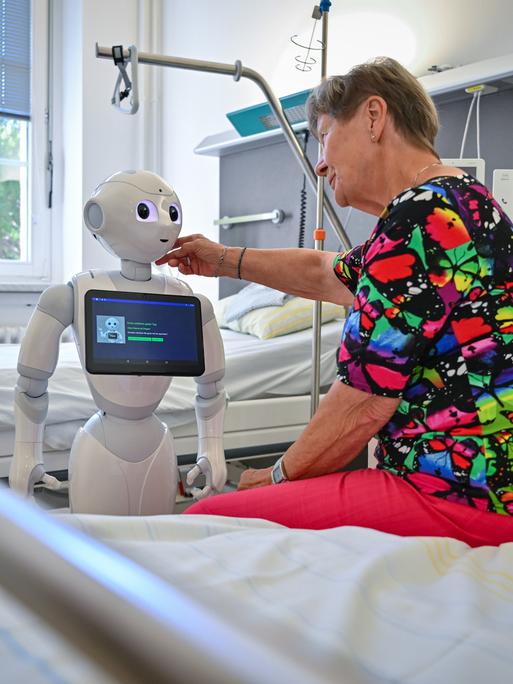 Eine ältere Dame sitzt auf einem Pflegebett und streicht einem humanoiden Roboter, der vor ihr steht, mit der rechten Hand über die Wange.