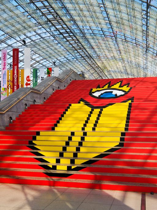 Das Logo der Leipziger Buchmesse auf einer Treppe in einer Glashalle der Messe in Leipzig. Auf rotem Grund ist ein stilisierter Kopf zu sehen, der aus nur einem Auge besteht. Davor, ebenfalls in Gelb, die Umrisse eines Buches. 