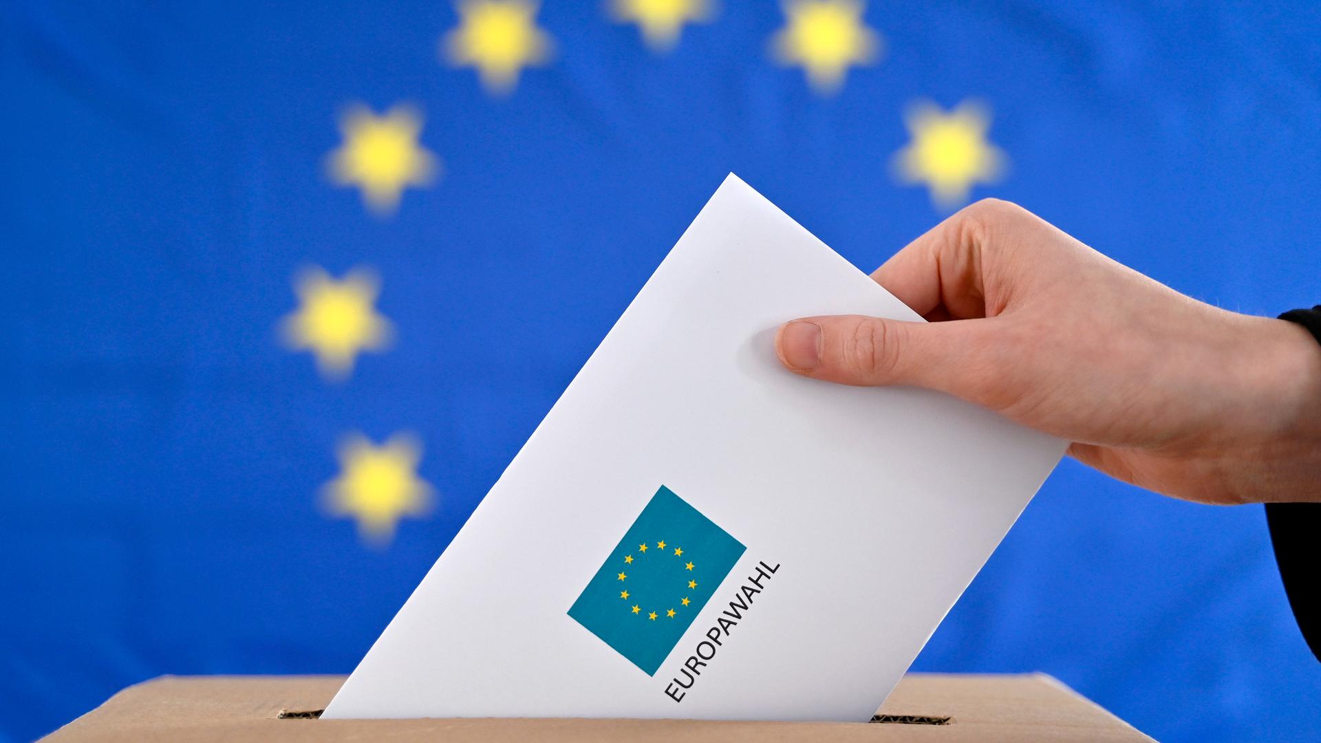 Ein Wahlbrief wird in eine Wahlurne gesteckt, im Hintergrund eine EU-Flagge.