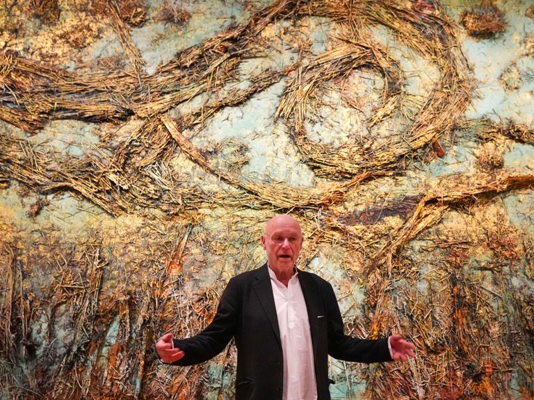 Anselm Kiefer steht vor einem seiner Werke im Van Gogh Museum in Amsterdam, blickt in die Kamera und breitet die Arme weit aus. 