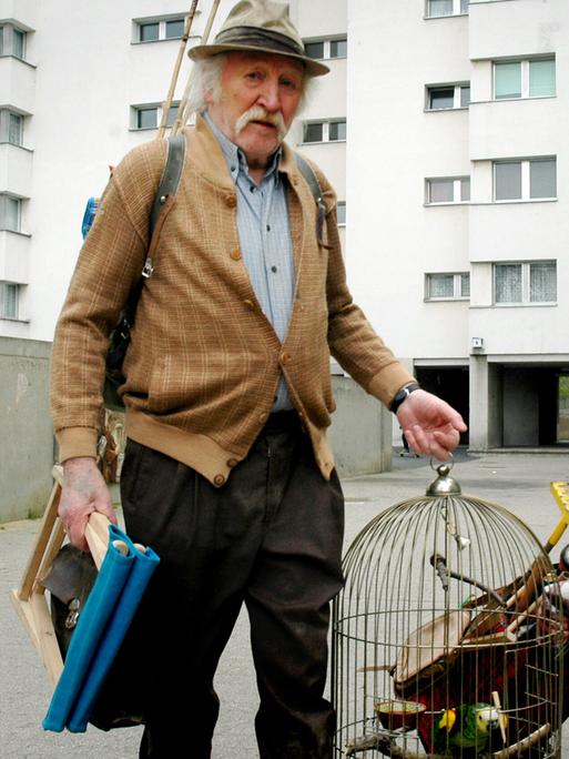 Umzug zu Fuß: Ein Mann mit Hut und weißem Schnauzbart trägt einen Vogelkäfig mit Papagei und seine Frau zieht einen Einkaufswagen hinter sich her, aus dem ein Rechen ragt.