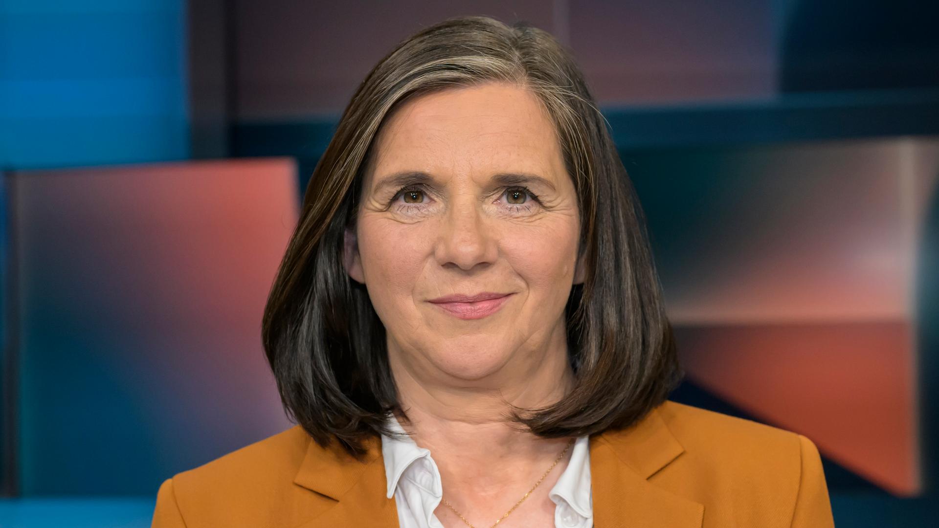 Die Bundestagsvizepräsidentin Katrin Göring-Eckardt (Bündnis 90/Die Grünen). Sie lächelt auf dem Porträtfoto verhalten. Sie trägt einen orangefarbenen Blazer, der Bild-Hintergrund ist unscharf. 