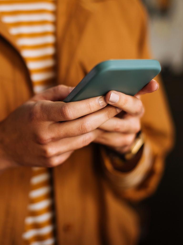 Nahaufnahme einer Hand, die ein Smartphone hält. Die dazugehörige Person trägt ein orangeweiß gestreiftes Shirt und eine orangefarbene Jacke.