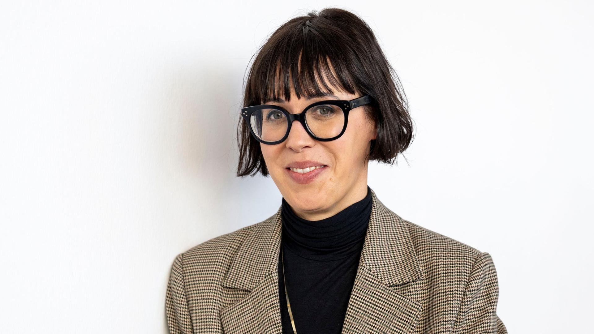 Eva Karl Faltermeier steht mit schwarzem Oberteil und kariertem Sakko vor einem weißen Hintergrund. Sie guckt durch eine schwarz umrandete Brille in die Kamera.