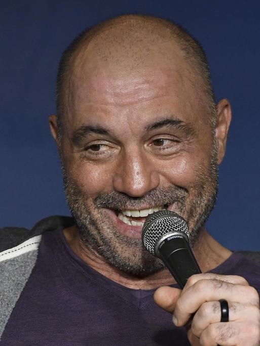 Das Bild zeigt den US-amerikanischen Comedia Joe Roegan, der mit seinem Podcast "The Joe Rogan Experience" berühmt geworden ist. Im Bild sitzt er mit kurzgeschorenen Haaren und breitem Lächeln mit einem Mikrofon in der Hand auf einer Bühne.