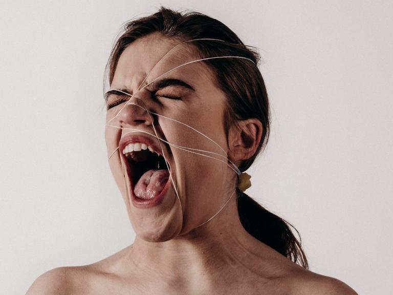 Eine Frau schreit mit weit geöffneten Mund, während ihr Gesicht von Schnüren eingesperrt wird.