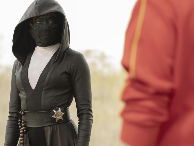 Eine schwarz verhüllte Frau mit Maske trägt einen Sheriffstern an der Hüfte.