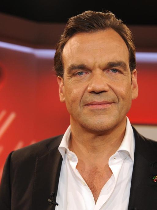 Christoph Gröner steht in einem rot gestalteten Studio und schaut freundlich in die Kamera