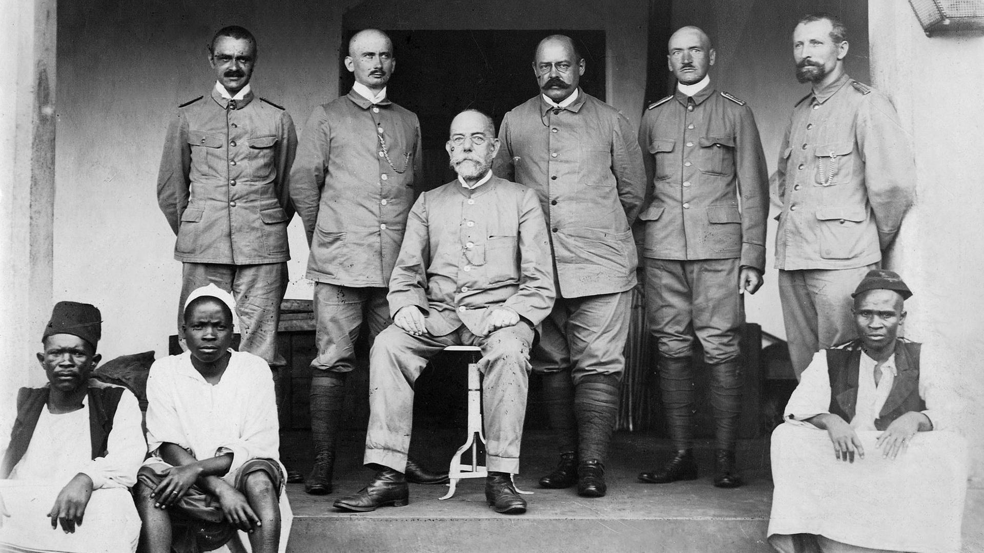 Robert Koch (sitzend) mit Mitgliedern der Expedition zur Untersuchung der Schlafkrankheit in Afrika; v.l.: Dr. Kudicke, Prof. Klein, Koch, Prof. Beck, Stabsarzt Pause, Feldwebel Sacher (Foto: Franz-Otto-Koch - 1897) Vor den Weißen hocken drei Schwarze auf den Stufen der Terrasse.