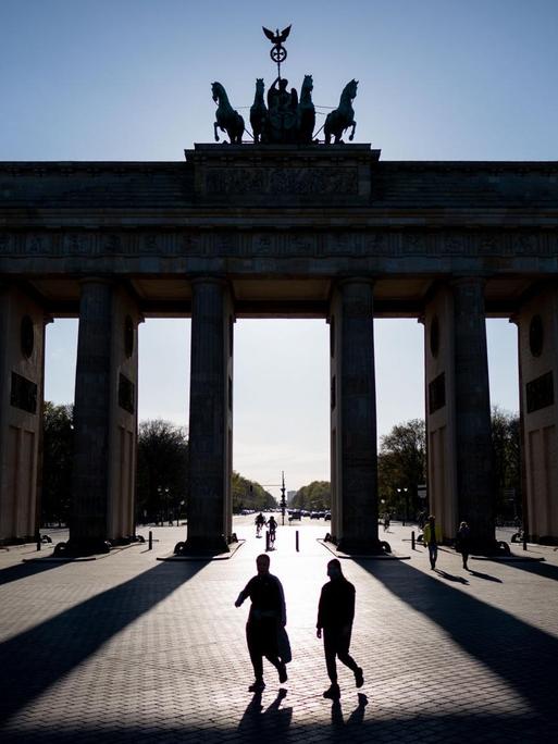 Das Brandenburger Tor in Berlin wirft in der untergehenden Sonne lange Schatten, zwei Personen gehen mittig davor entlang.