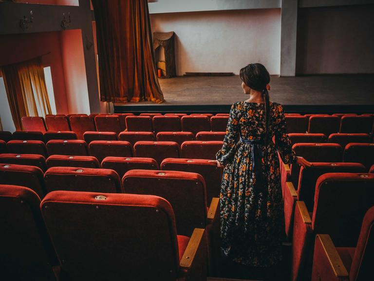 Im Theatersaal: Eine elegante Frau im Kleid steht im Gang zwischen den leeren Zuschauersitzen und blickt nachdenklich gen Theaterbühne.