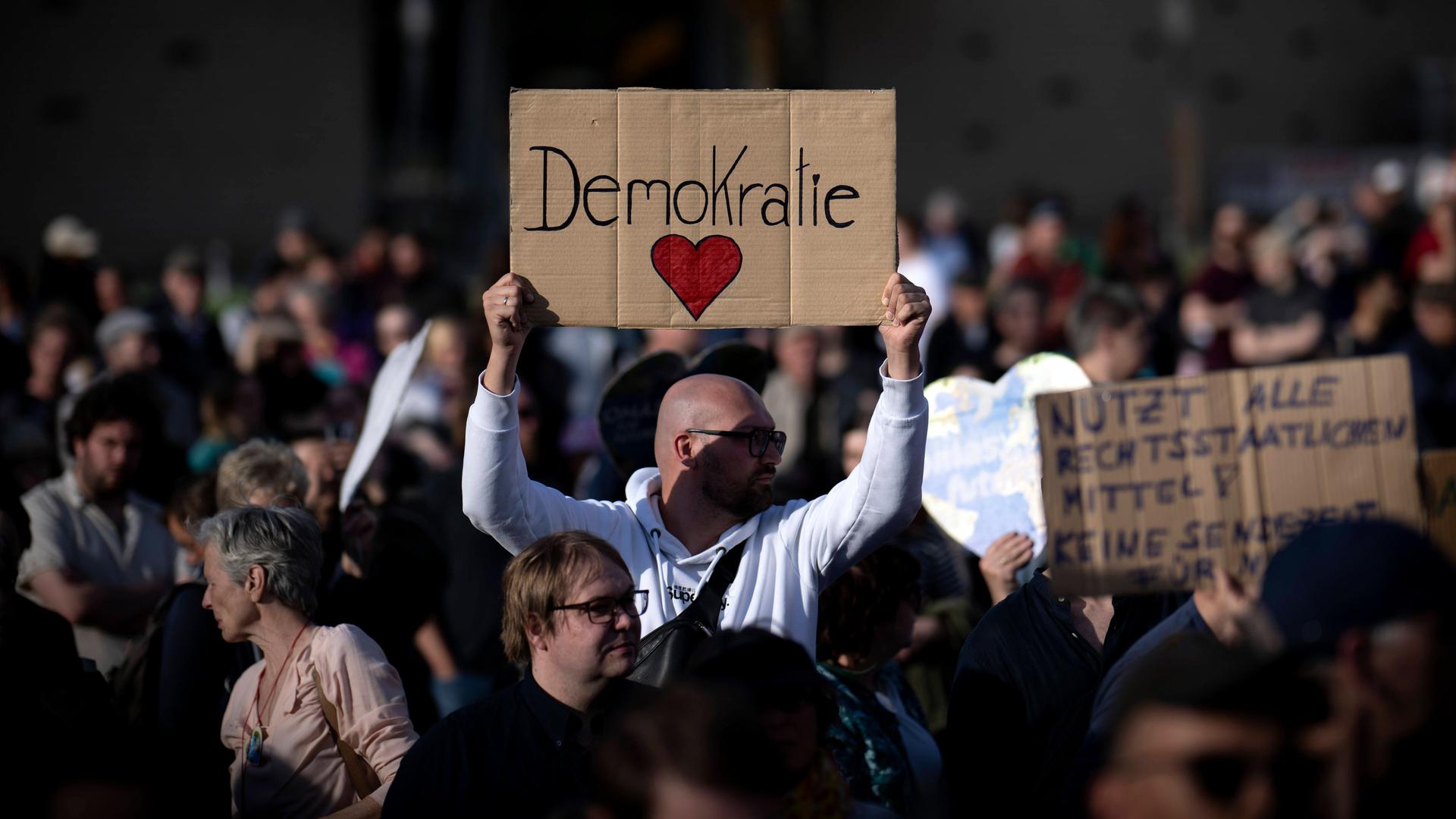 Ein Man hält auf einer Demo ein Schild in die Höhe, auf dem "Demokratie" steht. Darunter ist ein rotes Herz gemalt.