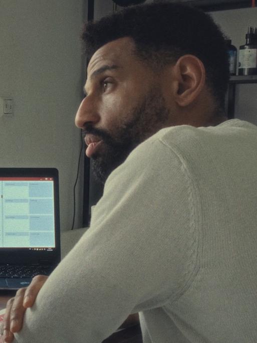 Der Protagonist aus dem Film „Soldaten des Lichts“ David (rechts im Bild) ist mit einem Anhänger seiner sektenähnlichen Gruppe im Gespräch. Die beiden sitzen am Schreibtisch. Im Hintergrund leuchtet ein Laptop.