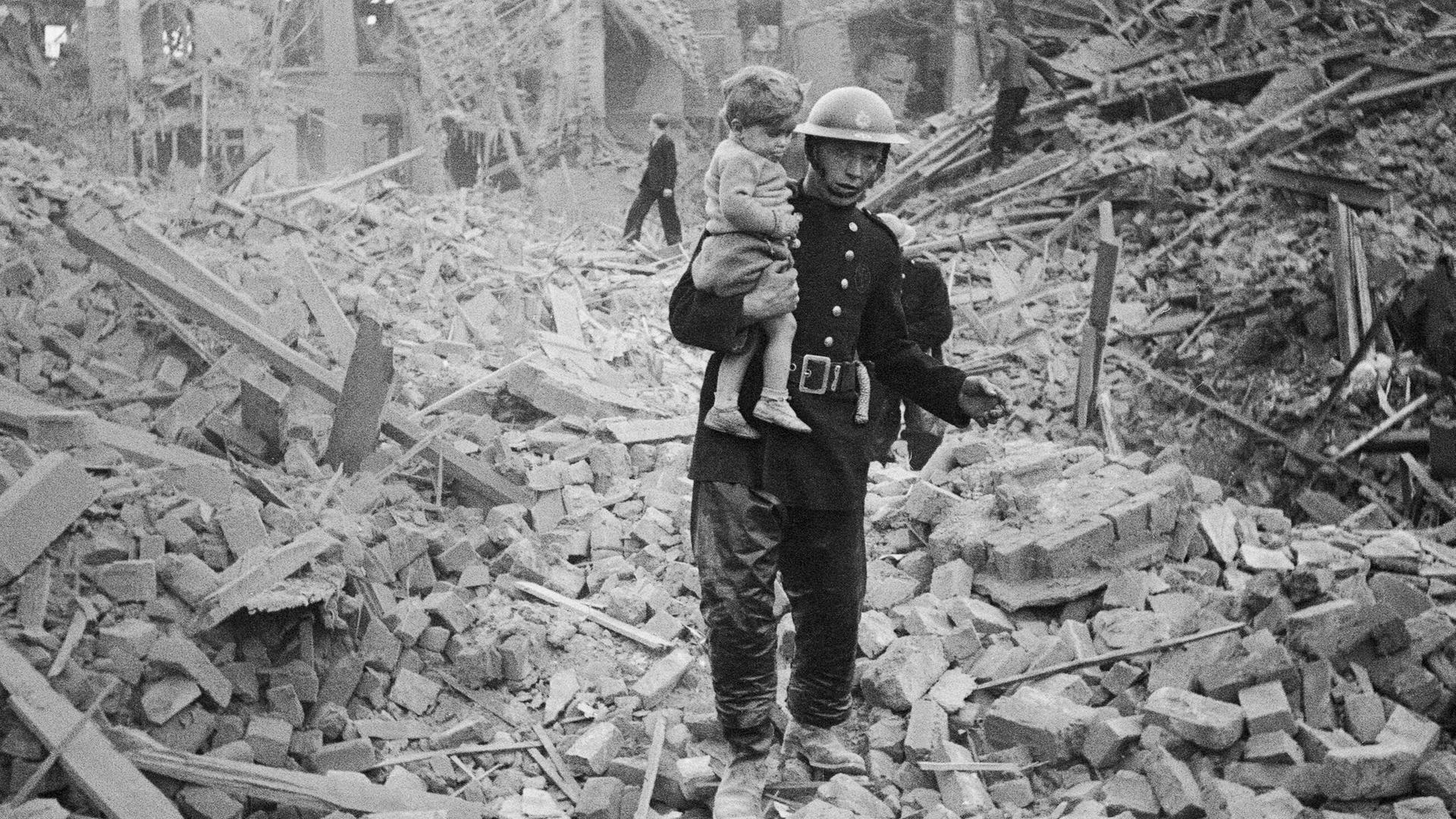 Ein Feuerwehrmann trägt ein Kleinkind nach einem Bombenangriff aus den Trümmern, London, um 1940. 