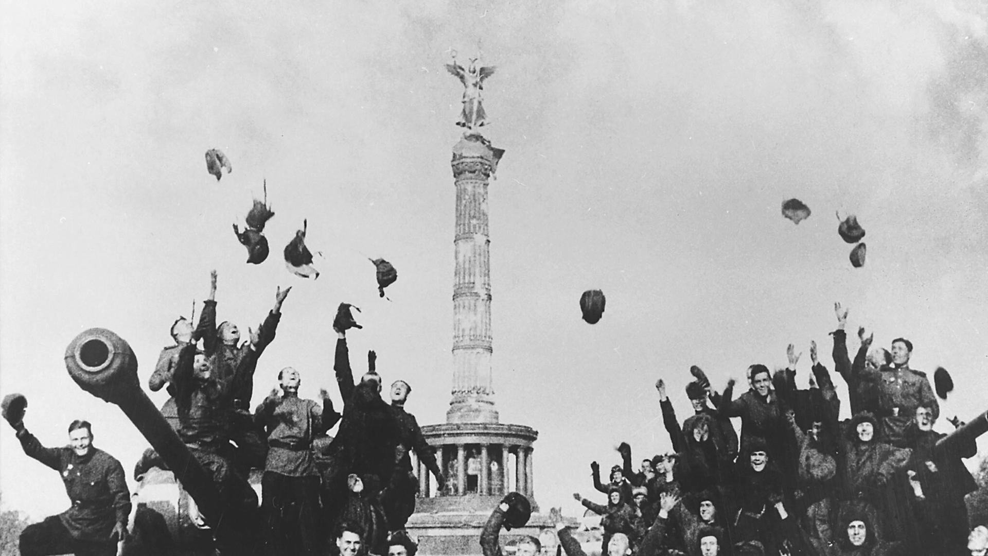 Historische Schwarz-Weiß-Aufnahme von sowjetischen Soldaten, die vor der Siegessäule in Berlin jubelnd ihre Helme und Hüte in die Luft werfen. Historische Schwarz-Weiß-Aufnahme von sowjetischen Soldaten, die vor der Siegessäule in Berlin jubelnd ihre Helme und Hüte in die Luft werfen.