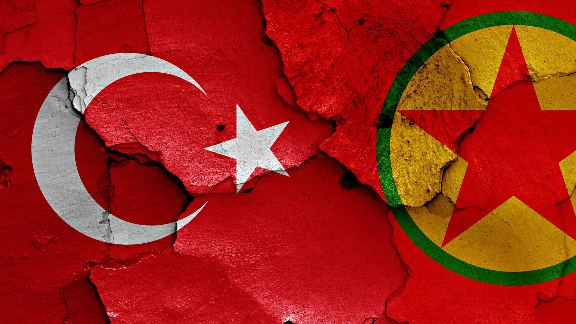 Ein türkischer Halbmond mit Stern und der kurdische rote Stern auf gelbem Kreis nebeneinander auf einer roten Fläche mit Rissen.