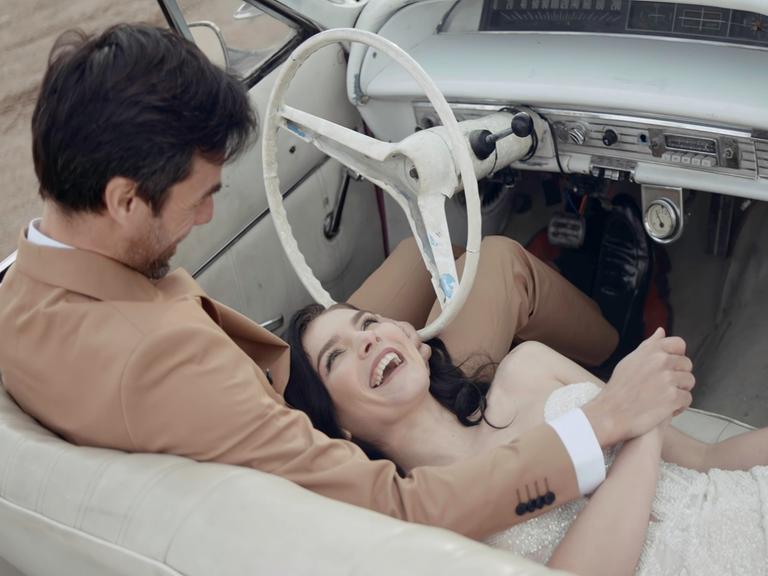 Glückliche lächelnde Frau, die in einem weißen Kleid, vermutlich einem Hochzeitskleid, auf den Knien ihres vermutlich frisch vermählten Mannes im Cabrio liegt. Er sitzt am Steuer und hält liebevoll ihre Hand.