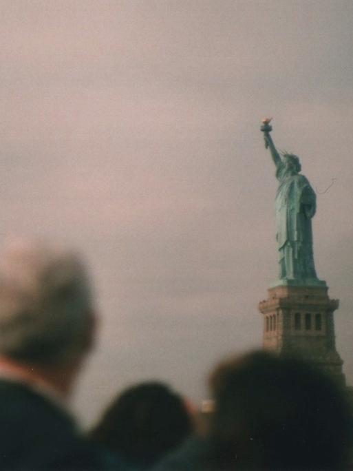 Menschen schauen auf die Freiheitsstatue, vor gräulich-flimmerndem Himmel.