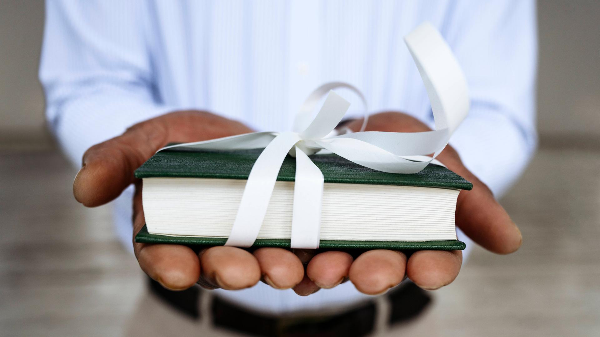 Nahaufnahme zweier Hände, die der Kamera ein mit weißem Geschenkband verziertes Buch entgegenhalten.