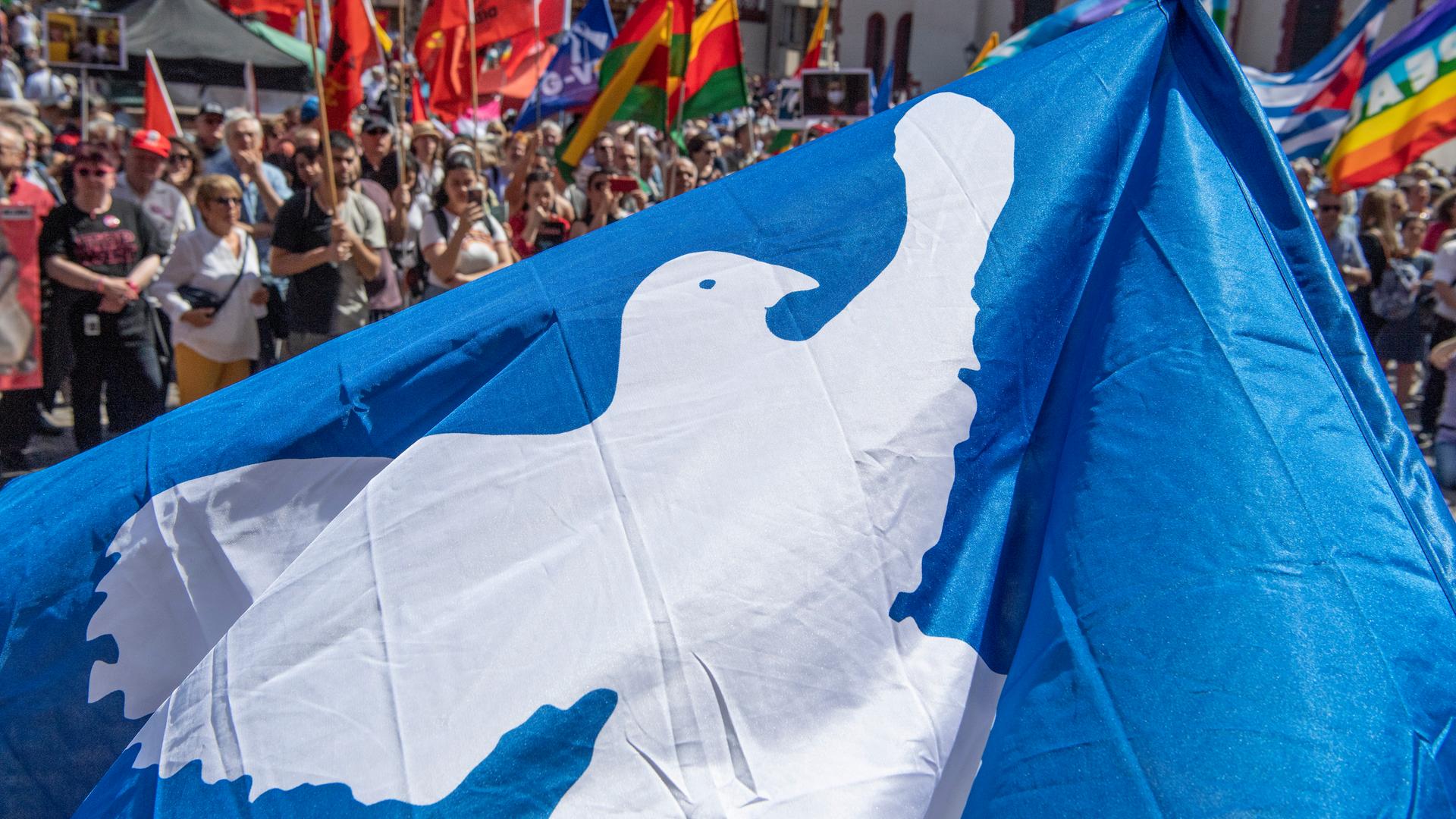 Auf einem Ostermarsch mit zahlreichen Teilnehmern ist eine blaue Fahne mit einer weißenn Friedenstaube zu sehen. 