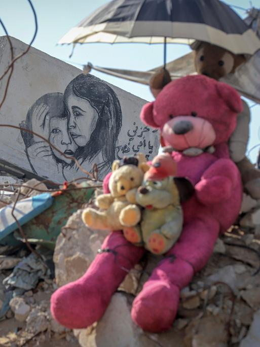 Teddybären sitzend auf einem Geröllhaufen eines eingestürzten Hauses in Gaza. Ein weiteres Stofftier hält einen kleinen Sonnenschirm über ihre Köpfe. Auf einem Stück Mauer ist eine Zeichnung auf der ein kleines Mädchen einen kleinen Jungen tröstend umarmt.