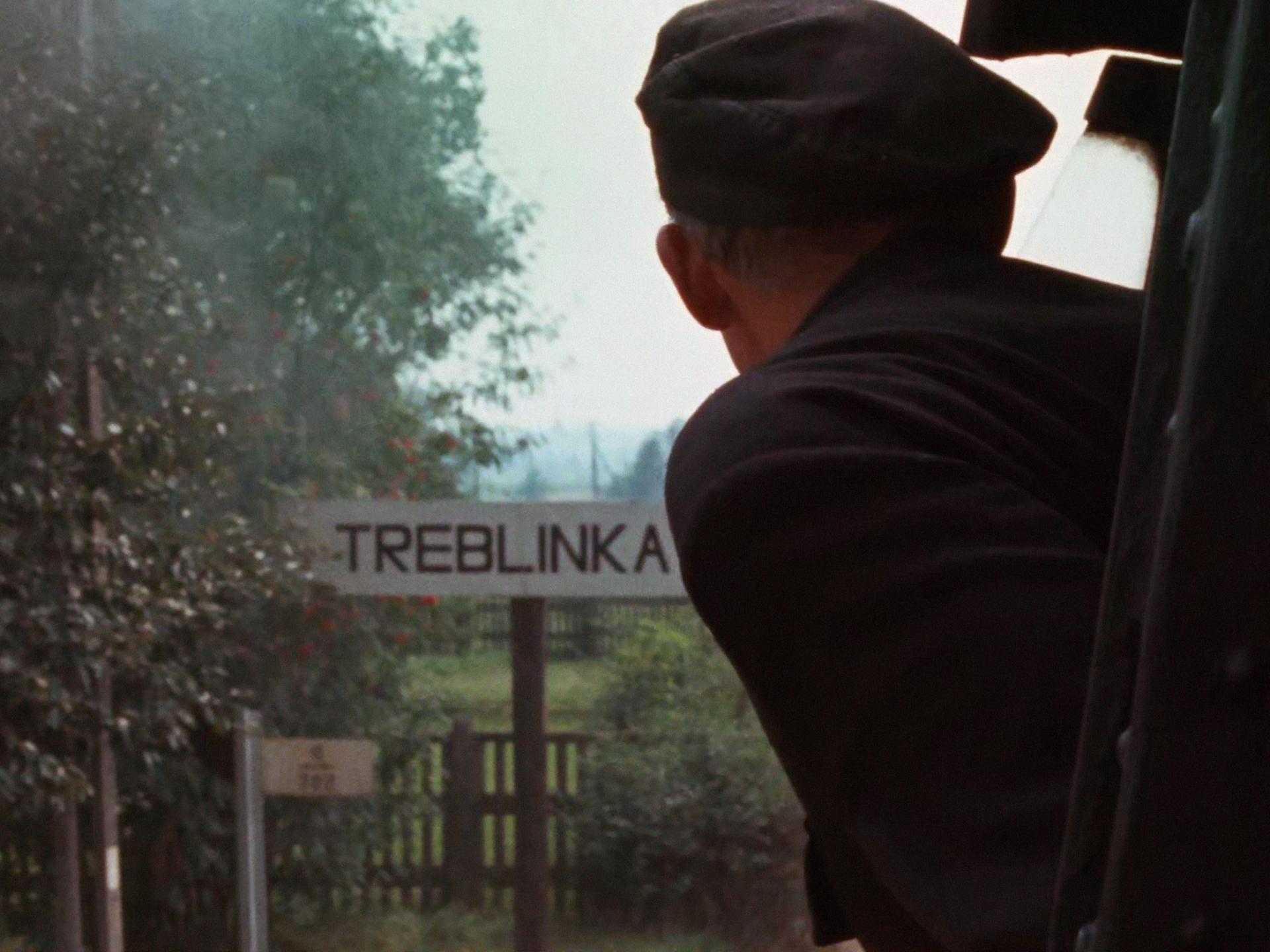 Szene aus Claude Lanzmanns Shoa. Eine Person guckt aus einem Zug, während dieser ein Schild mit der Aufschrift "Treblinka" passiert.