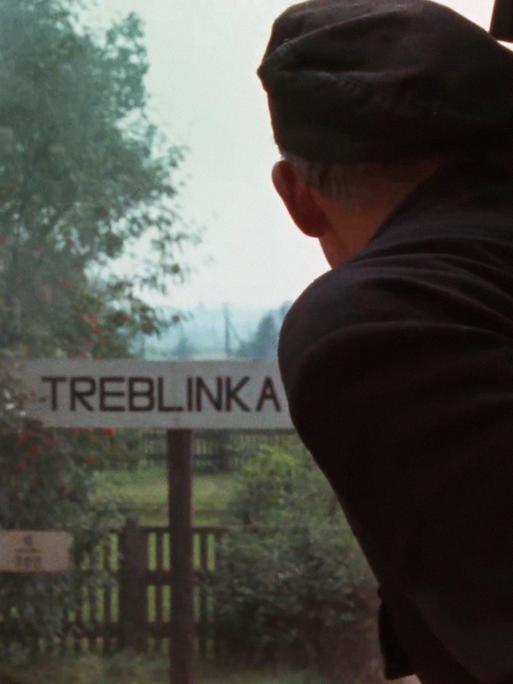 Szene aus Claude Lanzmanns Shoa. Eine Person guckt aus einem Zug, während dieser ein Schild mit der Aufschrift "Treblinka" passiert.