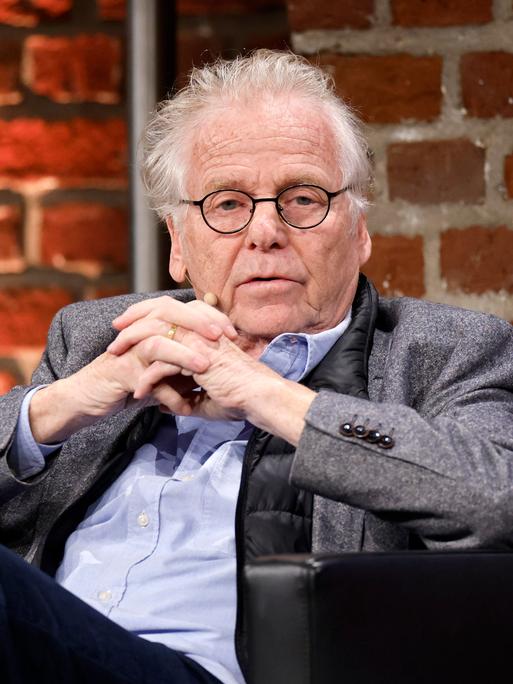 Der frühere Grünen-Politiker und Publizist Daniel Cohn-Bendit (Grüne) im grauen Jacket, hält die Hände verschränkt und sitzt vor einer Ziegelsteinwand. 