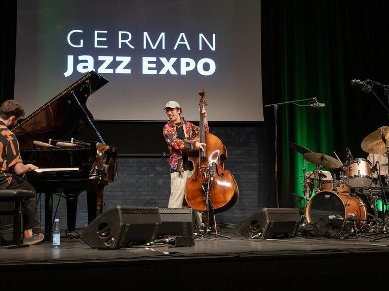 Der Bassist Nils Kugelmann steht im Zentrum einer Bühne auf der Messe jazzahead am Bass, neben ihm ein Flügel und ein Schlagzeug. 